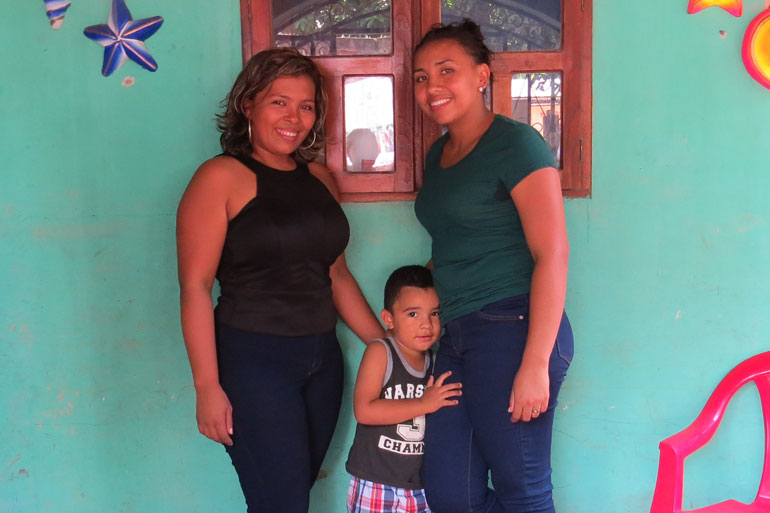 This image of two grown women and a young child standing in front of a brightly coloured wall. The two women are smiling and the child clutches one woman's leg with a small smile on his face.