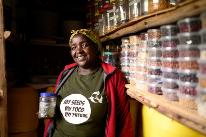 Beatrice Wangui stands proudly next to her shelf of colourful seeds in containers, wearing a shirt that says "my seeds, my food, my future"